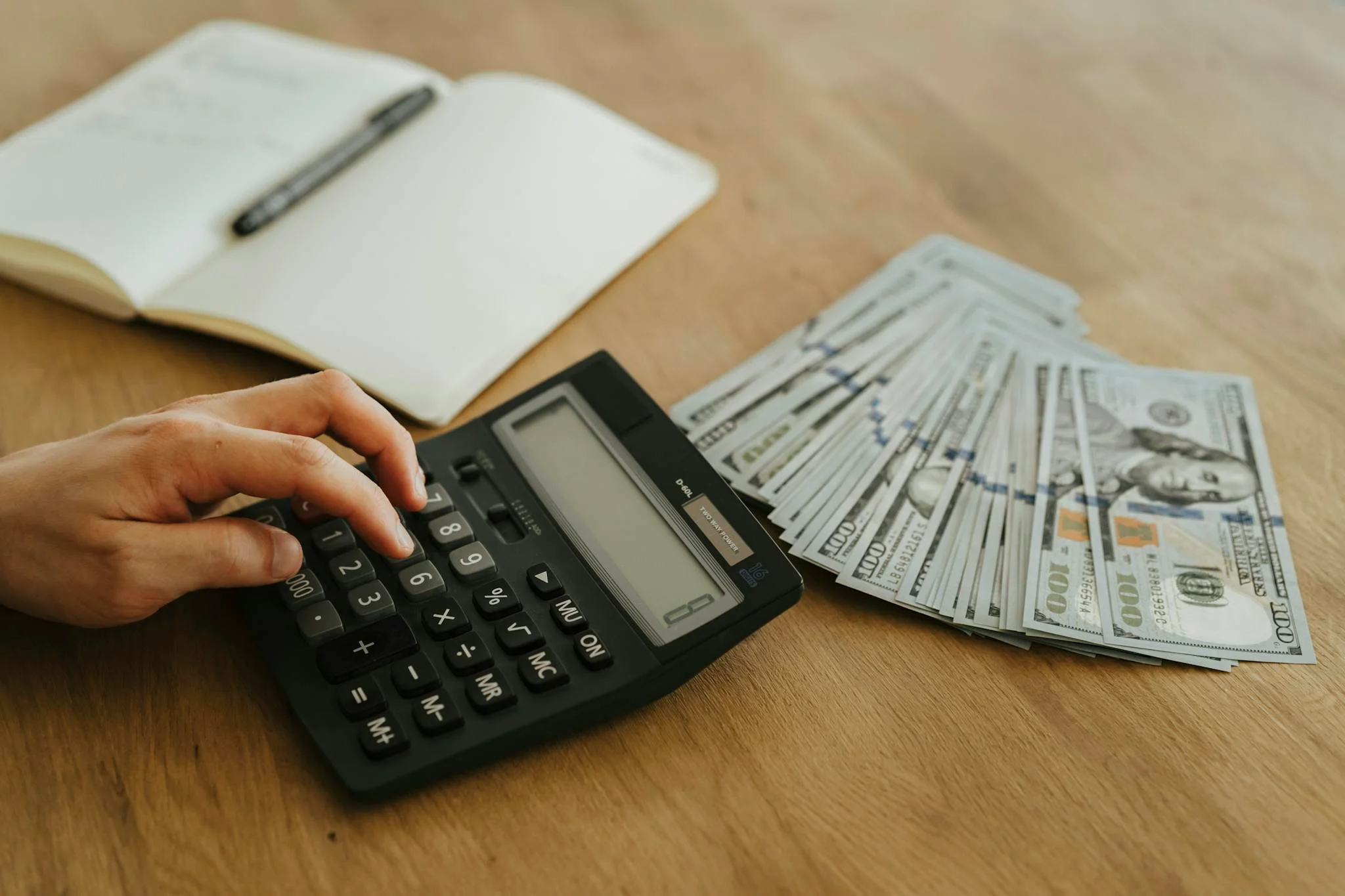 Close-up of a hand using a calculator with cash and a notebook on a wooden table.
